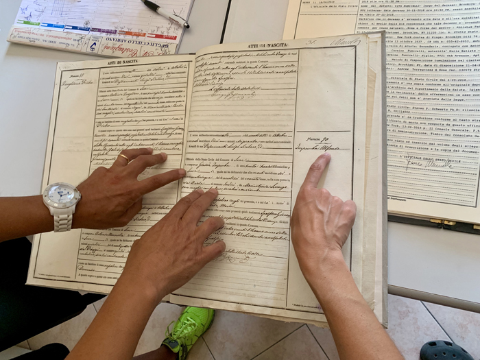 The hands of three people point to parts of an old handwritten ledger page