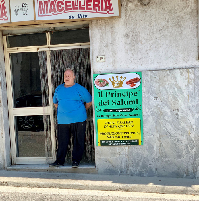 A man in a blue shirt poses in front of a store front. A sign, above, reads "Macelleria da Vito" and another sign advertises sausages.